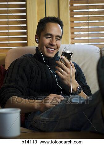 Stock Image - Man sitting on sofa with mp3 player listening to music smiling. Fotosearch