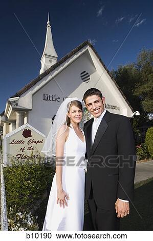 Stock Image - Portrait of a newlywed couple standing in front of a chapel and smiling. Fotosearch