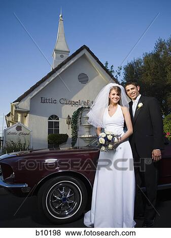 Stock Photo - Portrait of a newlywed couple standing near a car in front of a chapel. Fotosearch