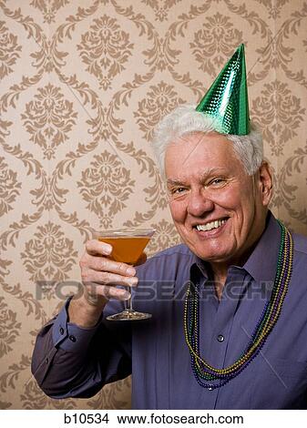 Picture - Portrait of a senior man holding a glass of martini and wearing a party hat. Fotosearch