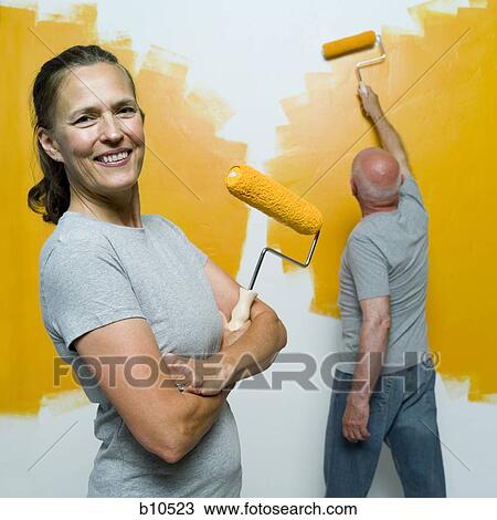 Stock Image - Portrait of a senior woman holding a paint roller with a senior man painting a wall. Fotosearch