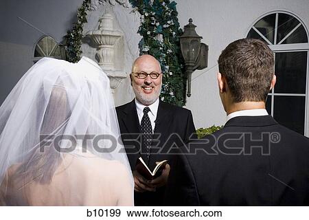 Stock Photo - Rear view of bride and groom in front of priest in a chapel. Fotosearch