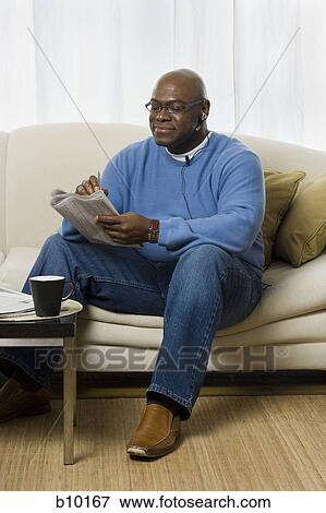 Senior man sitting on a couch and reading a newspaper View Large Photo Image Stock Photo - Senior man sitting on a couch and reading a newspaper. Fotosearch