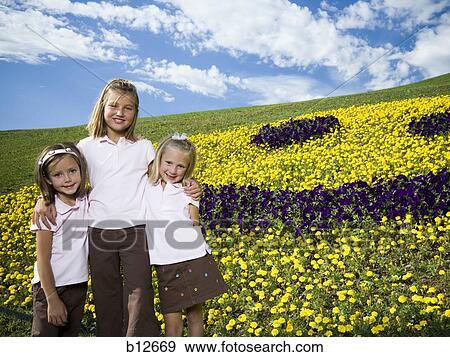 Stock Photo - Three sisters posing in front of happy face floral display. Fotosearch