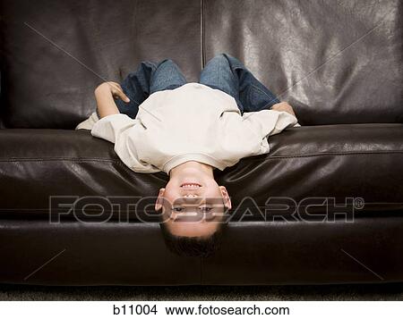 Picture - Young boy lying upside down on a leather sofa. Fotosearch