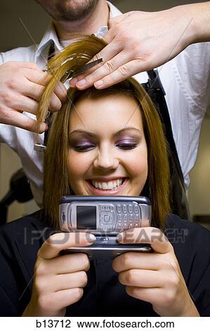 Stock Image - Young woman with camera phone having hair cut smiling. Fotosearch