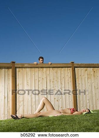 Stock Image - Man peeping over fence at woman sunbathing. Fotosearch