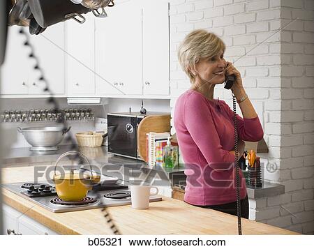 Stock Image - Mature woman talking on telephone in kitchen. Fotosearch
