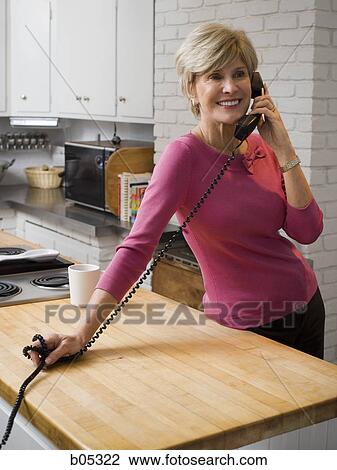 Stock Image - Mature woman talking on telephone in kitchen. Fotosearch
