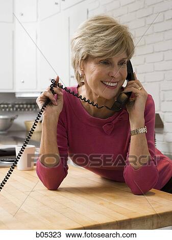 Stock Image - Mature woman talking on telephone in kitchen. Fotosearch