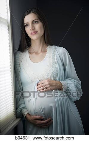 Stock Image - Pregnant woman in nightgown beside window. Fotosearch