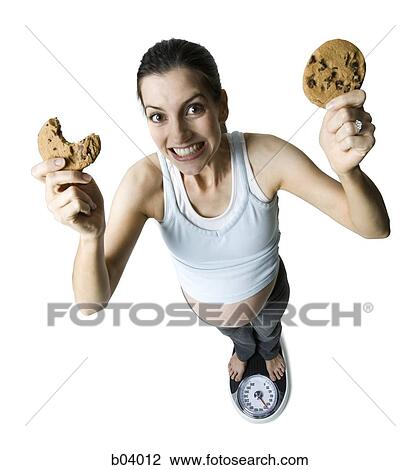 Stock Image - Pregnant woman on weigh scale with cookies. Fotosearch