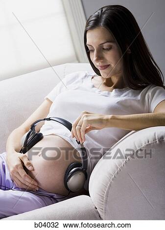 Stock Image - Pregnant woman sitting with headphones on belly. Fotosearch
