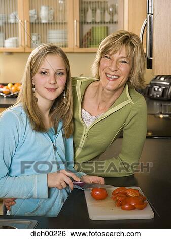 Stock Photograph - Portrait of a girl cutting tomatoes with her mother leaning against a kitchen counter. Fotosearch