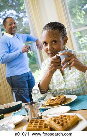 Stock Image - Portrait of a senior woman sitting at the breakfast table with a senior man standing behind her. Fotosearch