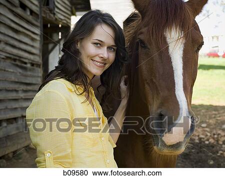 Stock Image - Portrait of a woman and a horse standing in front of a barn. Fotosearch
