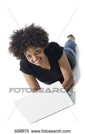 Stock Image - Portrait of a young woman lying on the floor and using a laptop. Fotosearch