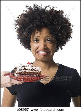 Stock Photo - Portrait of a young woman making a face with a person holding a cake in front of her. Fotosearch