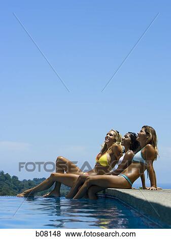 Stock Photo - Three women sitting by edge of infinity pool. Fotosearch