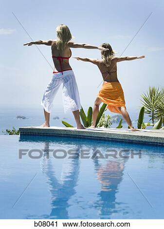 Stock Image - Two women performing yoga exercises by infinity pool. Fotosearch