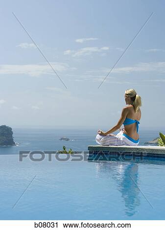 Stock Image - Woman performng yoga exercises by infinity pool. Fotosearch