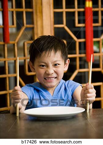 Boy sitting at table with empty plate holding chopsticks scowling View Large Photo Image Stock Photograph - Boy sitting at table with empty plate holding chopsticks scowling. Fotosearch