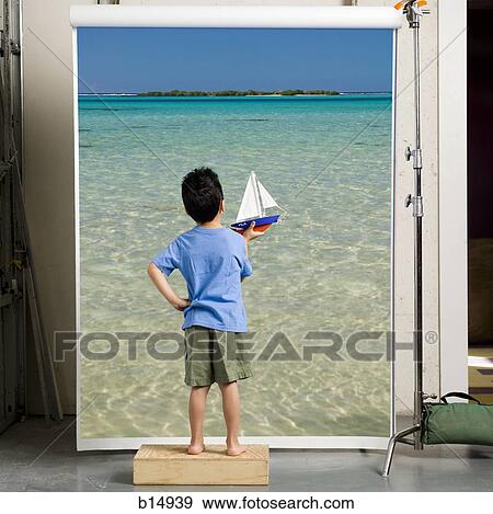 Stock Photo - Boy with toy boat looking at ocean rear view. Fotosearch