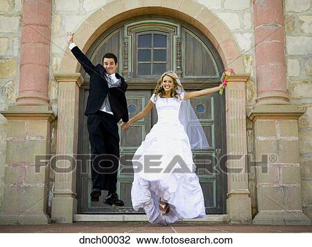 Stock Image - Bride and groom jumping and holding hands outside with bouquet. Fotosearch