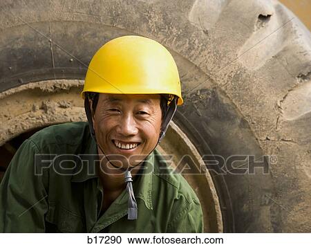 Stock Image - Construction worker sitting on tire of large machine smiling. Fotosearch