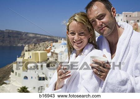 Stock Image - Couple in bath robes with coffee cups embracing and smiling with village in background. Fotosearch