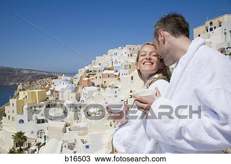 Stock Image - Couple in bathrobes with cups smiling with scenic village in background. Fotosearch