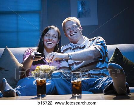 Stock Image - Couple sitting on sofa watching television with bowl of popcorn smiling. Fotosearch