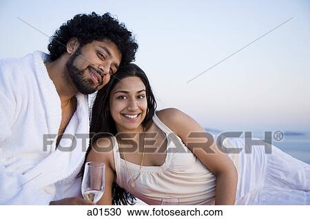 Stock Image - Couple sitting outdoors with champagne flutes and scenic background smiling and snuggling. Fotosearch