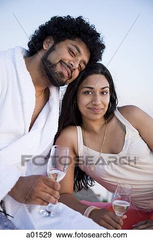 Stock Photo - Couple sitting outdoors with champagne flutes and scenic background smiling and snuggling. Fotosearch