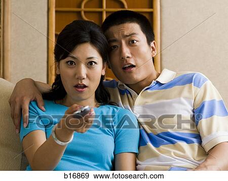 Couple watching television looking shocked with woman holding remote View Large Photo Image Stock Photo - Couple watching television looking shocked with woman holding remote. Fotosearch