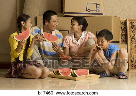 Stock Image - Family of four eating watermelon and smiling with cardboard boxes in background. Fotosearch
