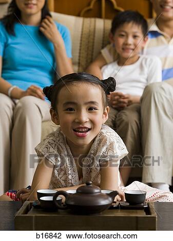 Stock Image - Girl in living room with tea pot and cups with parents and brother in background. Fotosearch