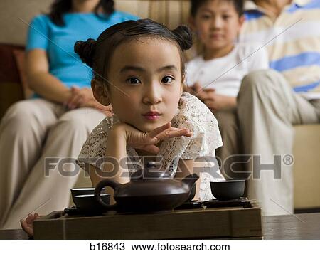 Stock Image - Girl in living room with tea pot and cups with parents and brother in background. Fotosearch