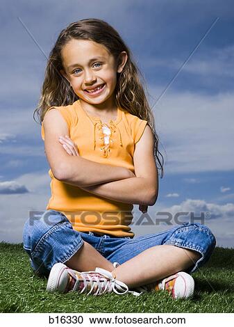 Stock Image - Girl sitting cross legged on grass outdoors with arms crossed smiling. Fotosearch