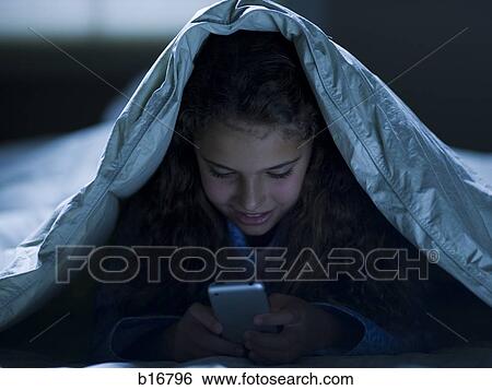 Stock Photograph - Girl under blankets at night with a smart phone. Fotosearch