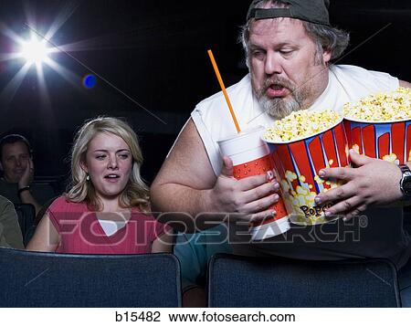 Stock Image - Large man with buckets of popcorn and drink at movie theater. Fotosearch