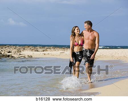 Stock Photo - Man and woman walking on shoreline embracing. Fotosearch