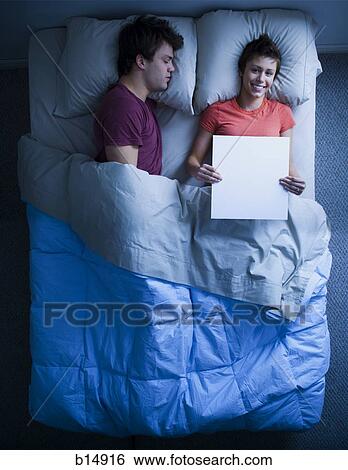 Stock Photograph - Man asleep in bed and woman holding blank sign smiling. Fotosearch