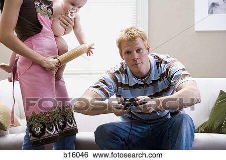 Man on sofa with video game controller and woman in apron with rolling pin holding baby View Large Photo Image Stock Photograph - Man on sofa with video game controller and woman in apron with rolling pin holding baby. Fotosearch