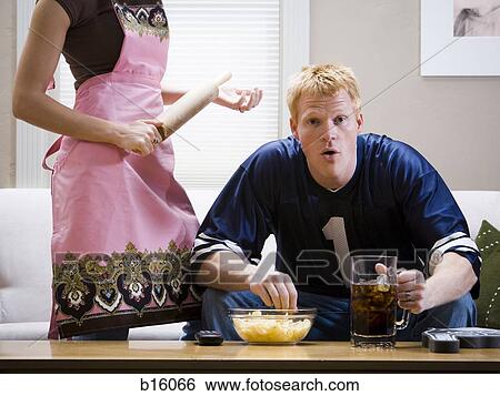 Stock Photograph - Man sitting on sofa watching television with beverage and chips with woman standing in apron with rolling pin. Fotosearch
