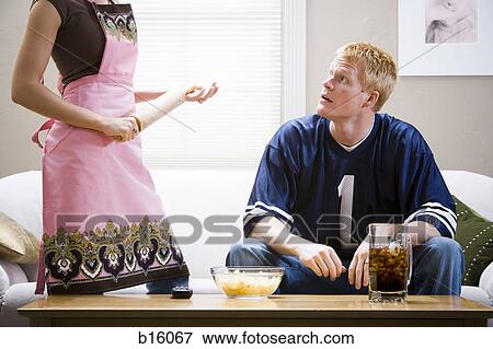 Stock Photo - Man sitting on sofa with beverage and chips with woman standing in apron with rolling pin. Fotosearch