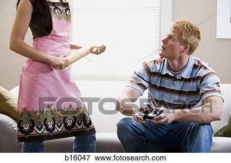 Man sitting on sofa with video game controller and woman with rolling pin in apron standing View Large Photo Image Stock Photo - Man sitting on sofa with video game controller and woman with rolling pin in apron standing. Fotosearch