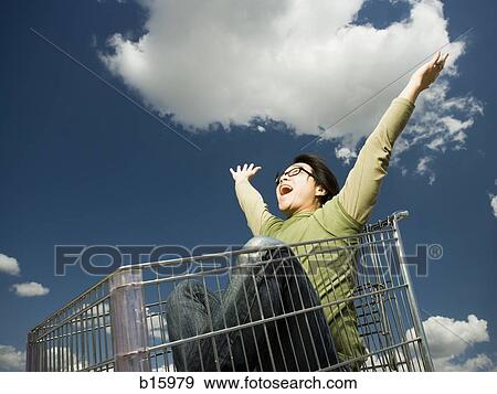Stock Photo - Man with eyeglasses in shopping cart outdoors with arms up smiling. Fotosearch