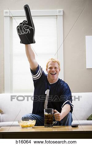 Man with giant glove and beverage sitting on sofa smiling and cheering View Large Photo Image Picture - Man with giant glove and beverage sitting on sofa smiling and cheering. Fotosearch