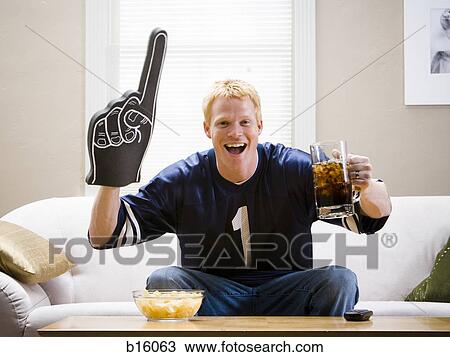 Man with giant glove and beverage sitting on sofa smiling and cheering View Large Photo Image Stock Image - Man with giant glove and beverage sitting on sofa smiling and cheering. Fotosearch
